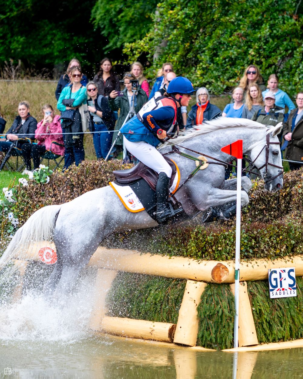 Eventing rider on a grey horse jumping into a water complex during a cross-country round, water splashing as spectators watch from behind.