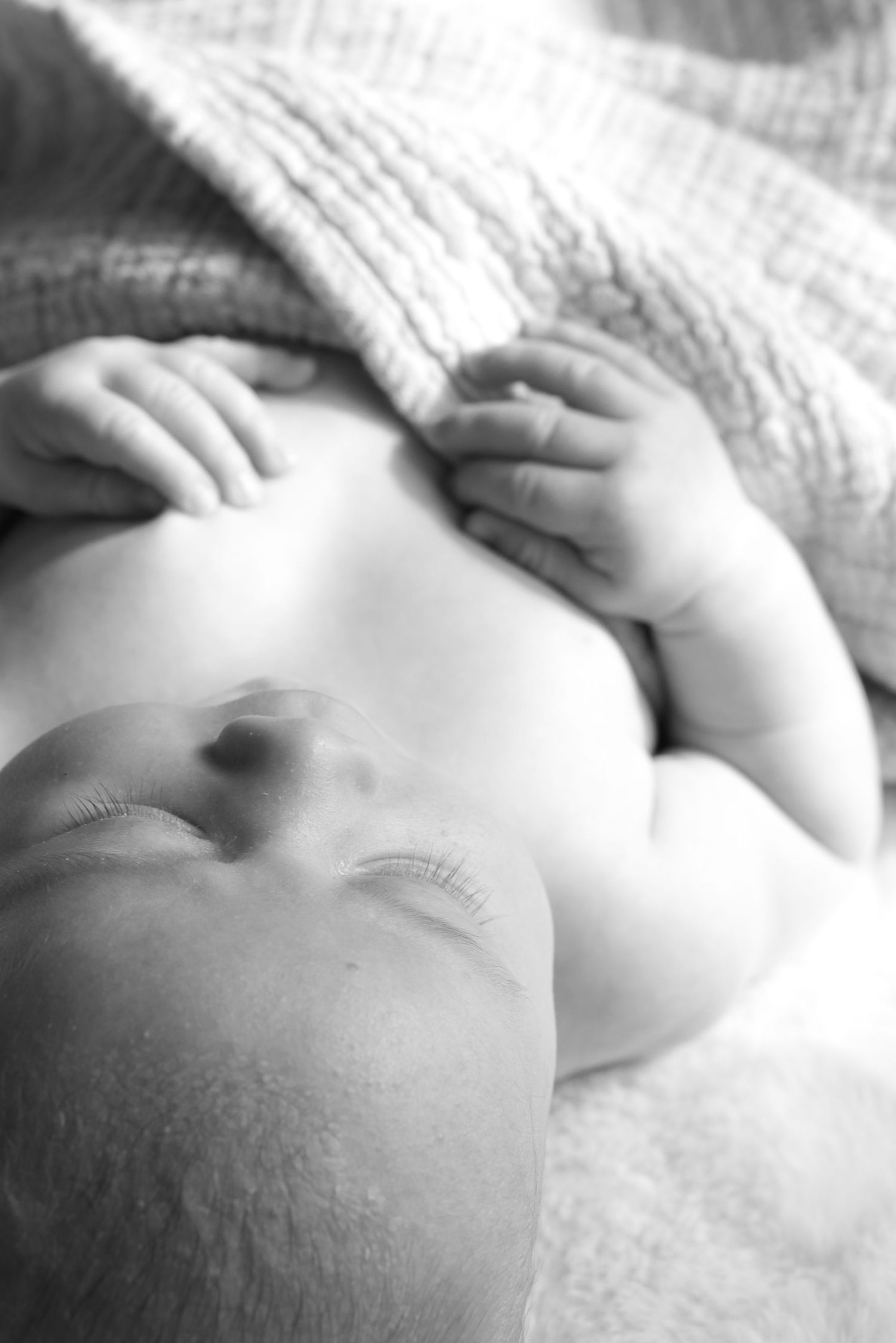A black and white image of a newborn baby. Taken from the head of the baby, looking down. The baby is sleeping and has a blanket draped over