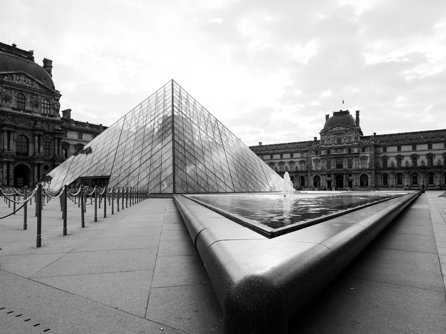 Black and white architectural photo of the glass pyramid entrance at the Louvre Museum in Paris with historic building facade.