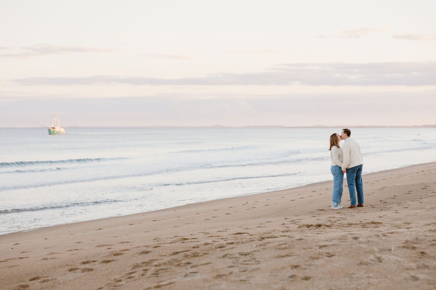 Barefoot couple standing close together on sandy beach with ocean waves in background.