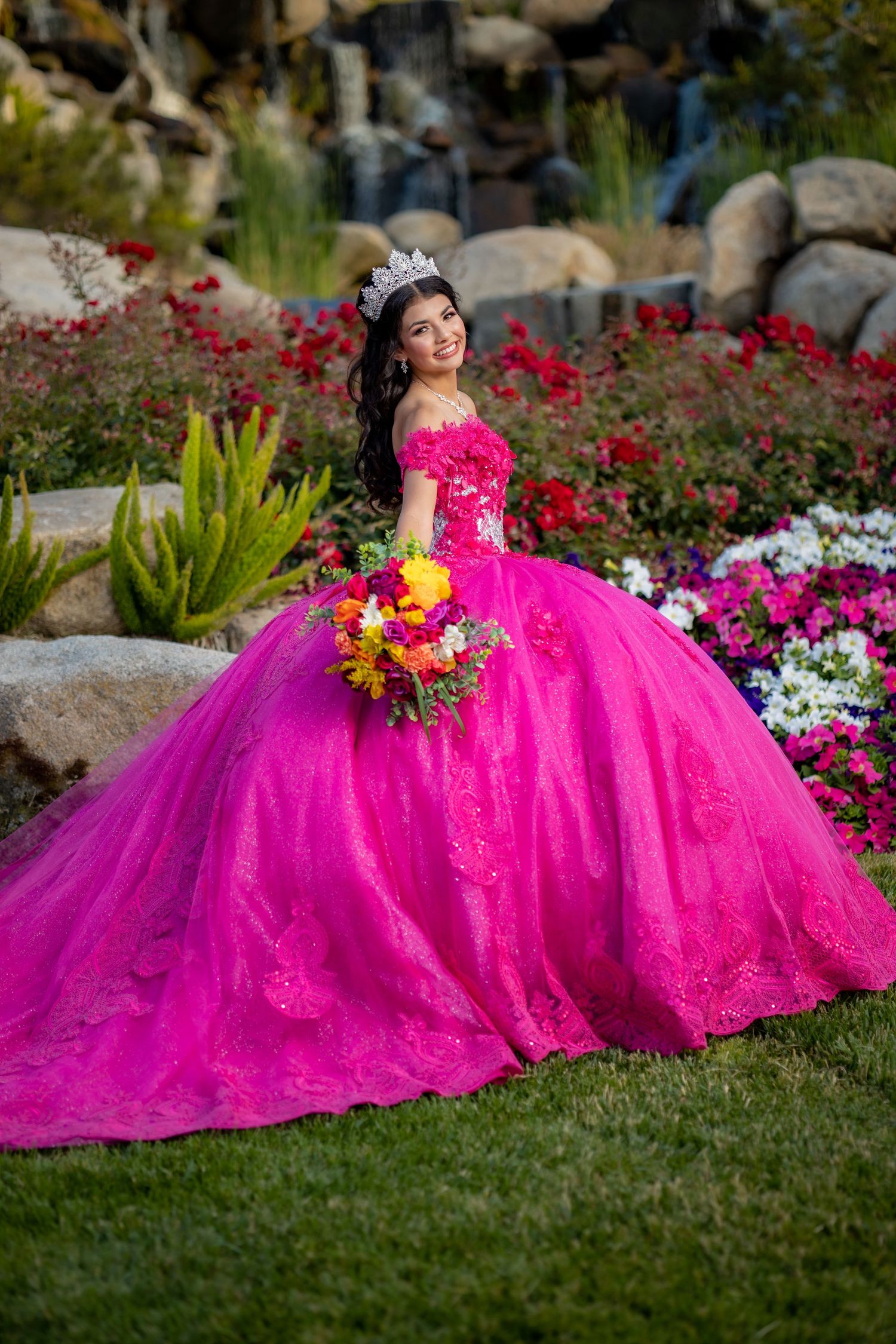Young woman in hot pink quinceañera ball gown with crown holds bouquet in a vibrant flower garden.