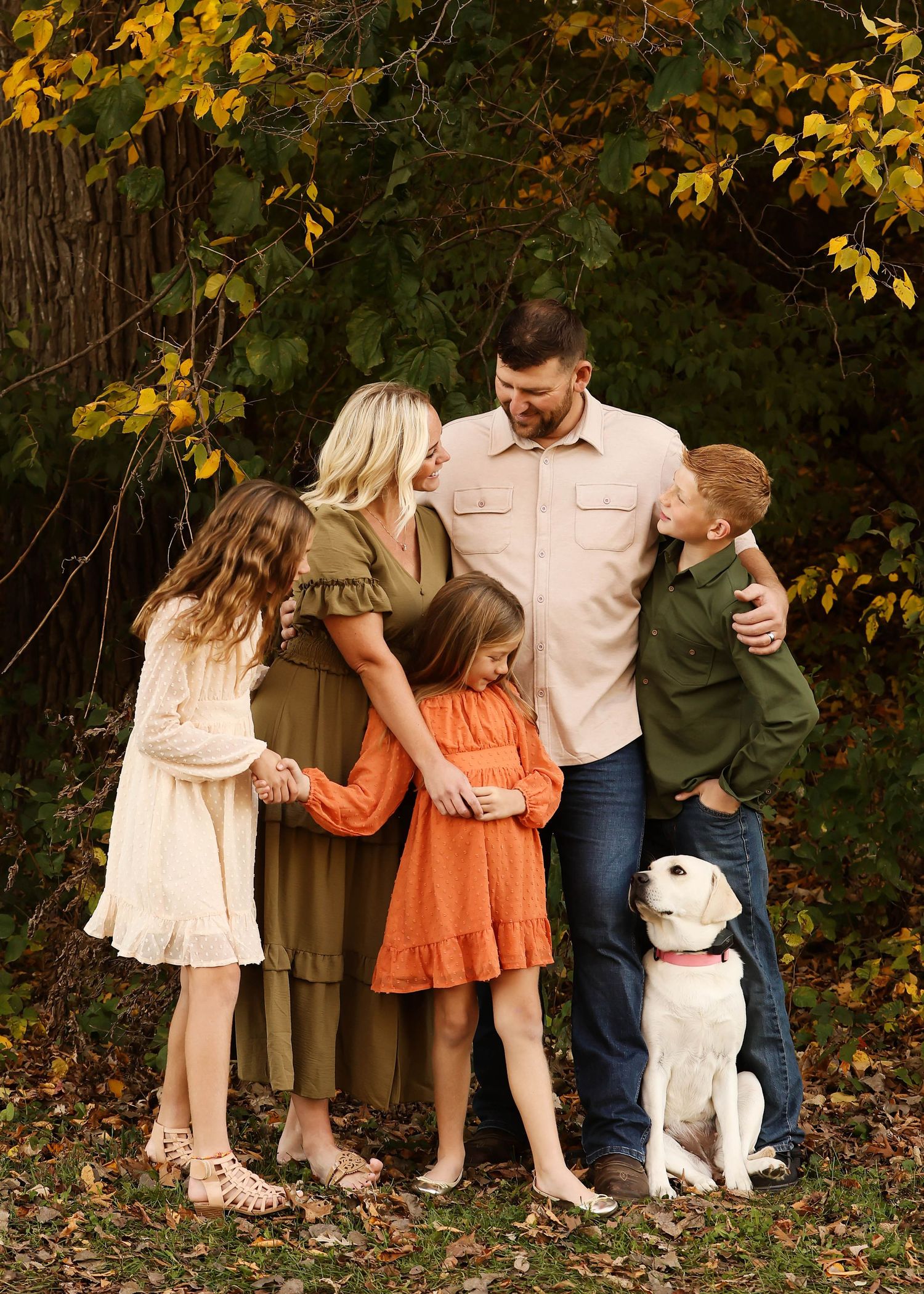 Family with dog poses playfully against autumn trees with golden leaves.