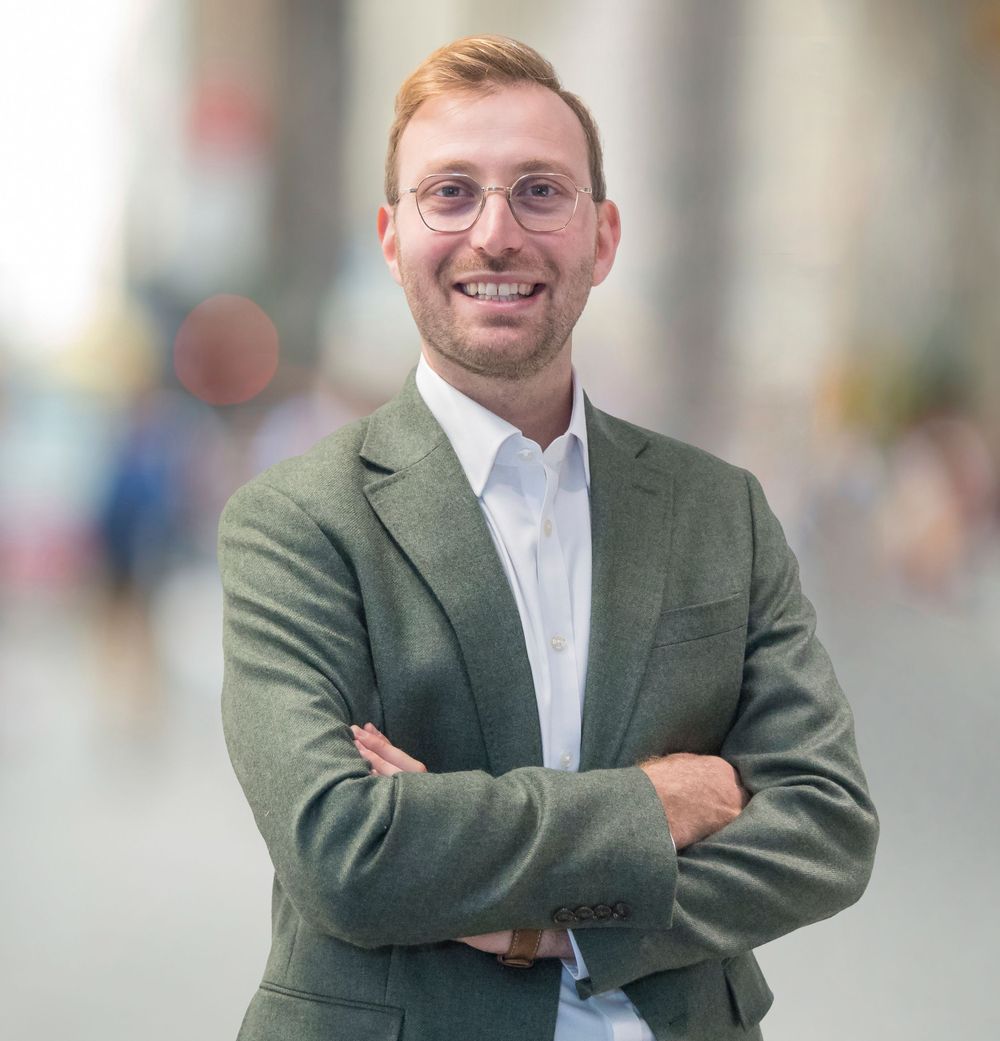 A professional headshot of a person in business attire with arms crossed in an urban setting.