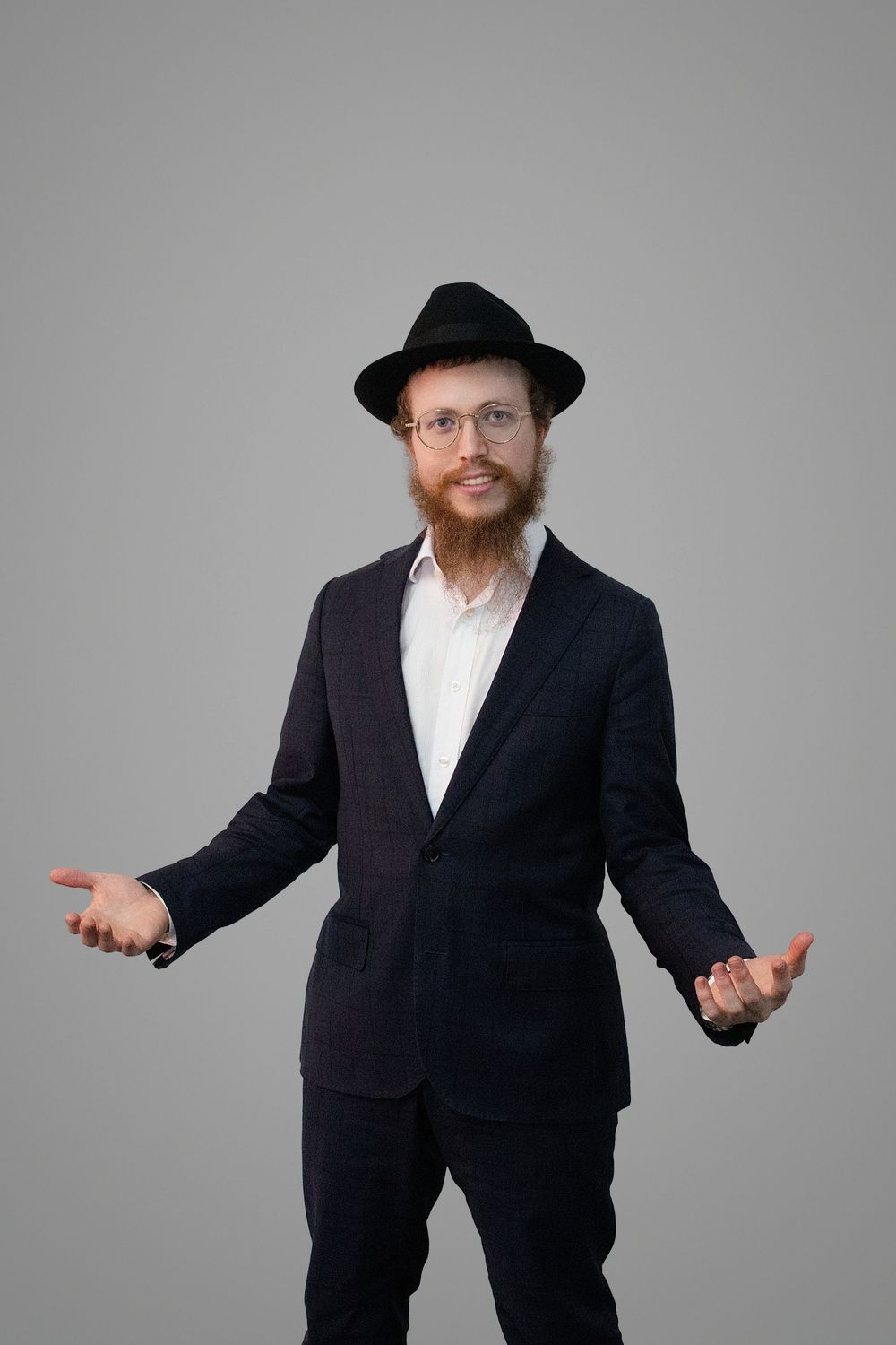 A person in a black suit and hat gestures with outstretched arms against a gray background.