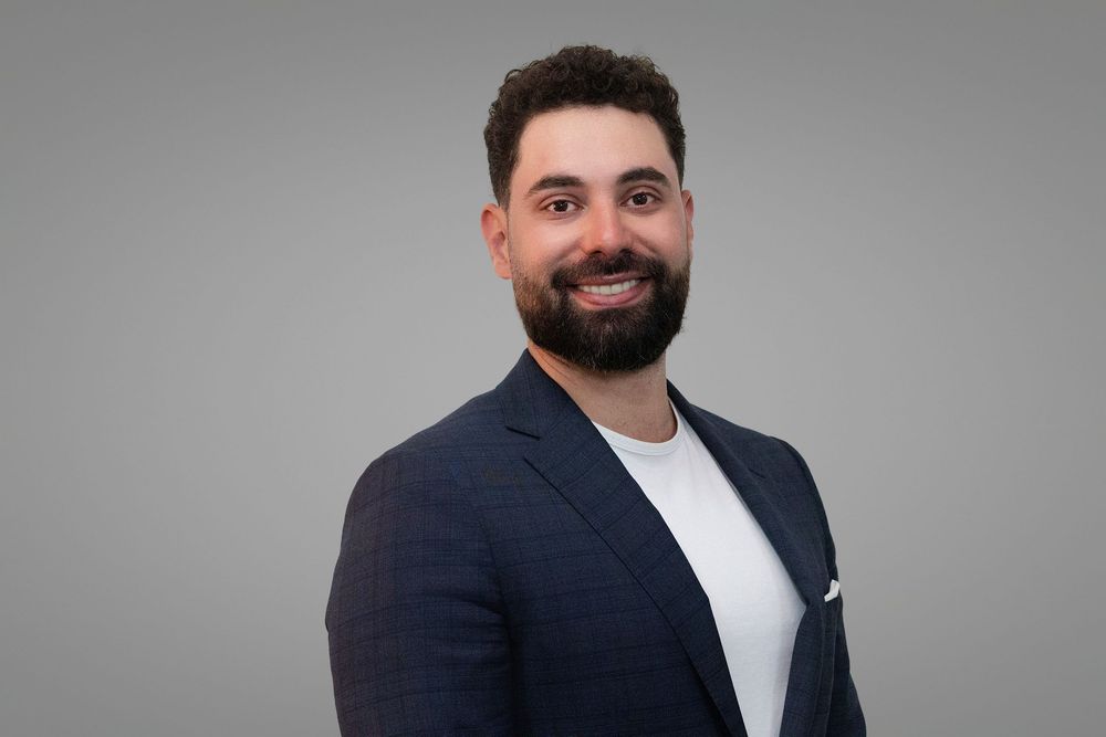 Professional headshot of someone wearing a white shirt and navy blazer against a gray background.