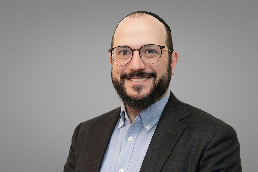 Professional headshots of a person wearing glasses and a dark suit against a gray background.