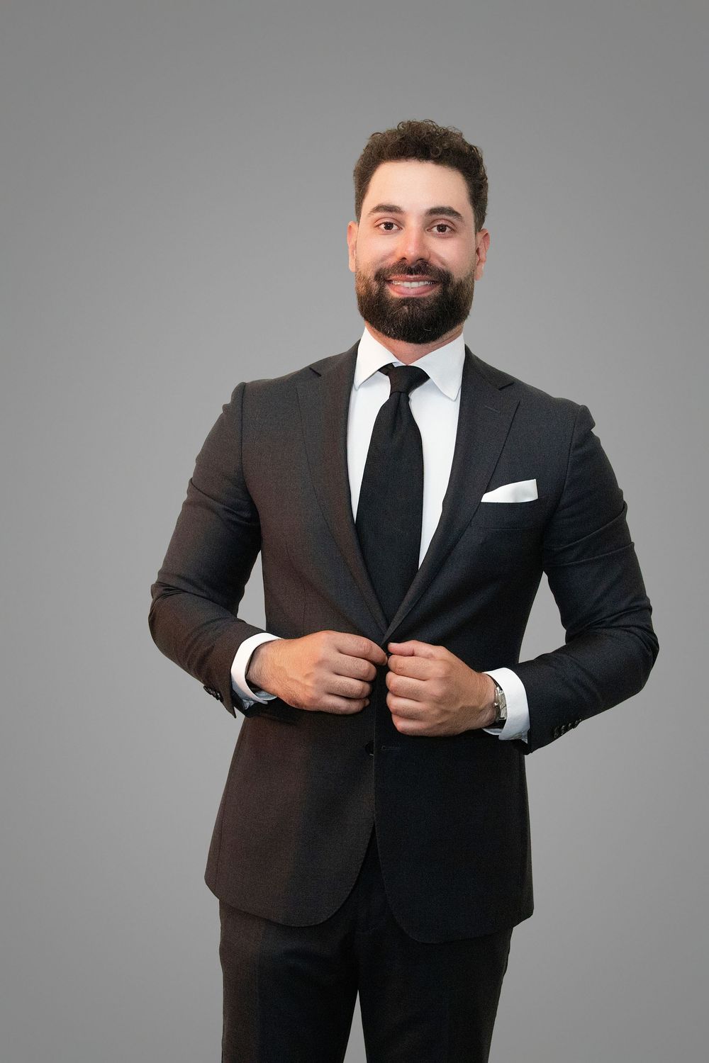Professional wearing a black suit and tie adjusts his jacket while posing in a studio setting against a gray background.