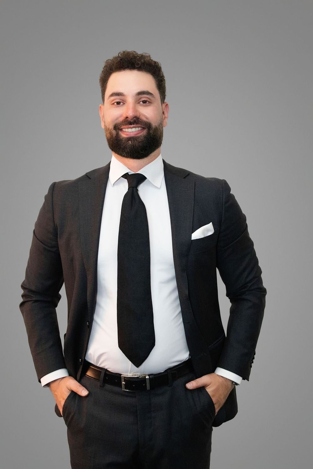 Professional portrait of a bearded person in a dark business suit with black tie and white pocket square against gray background.