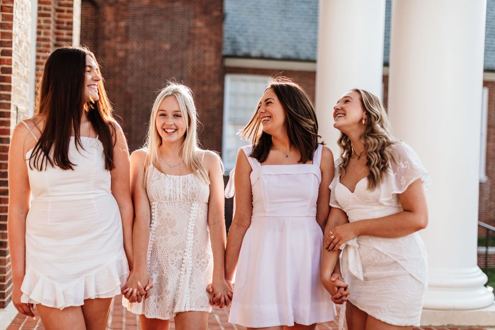 Four women holding hands and laughing together while wearing coordinated white summer dresses.