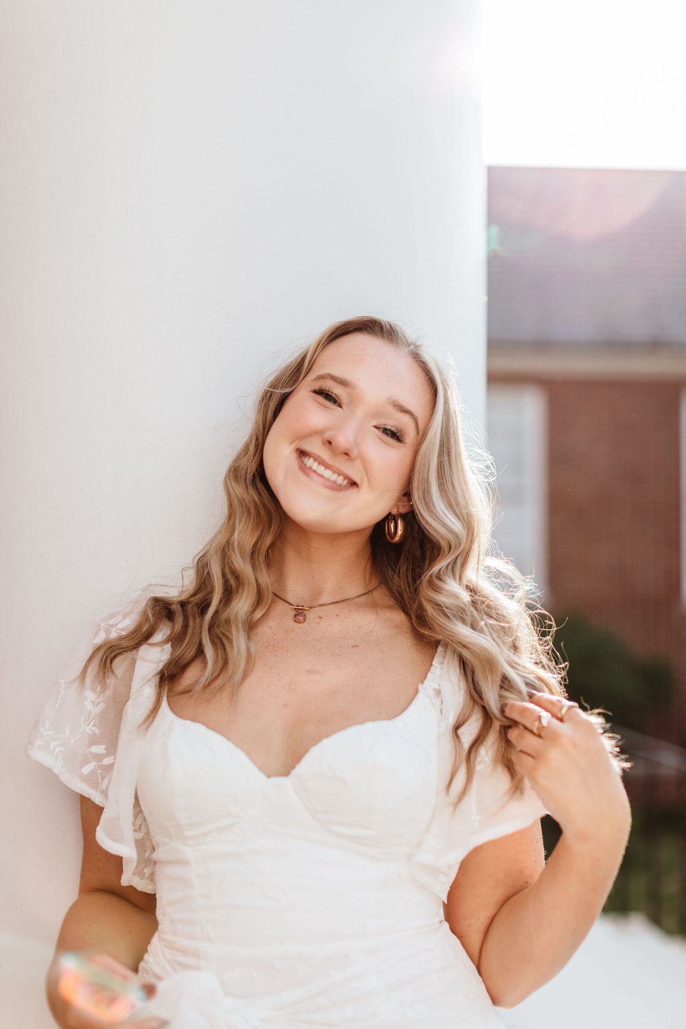 A smiling person in a white dress with wavy blonde hair wearing hoop earrings and a delicate necklace.