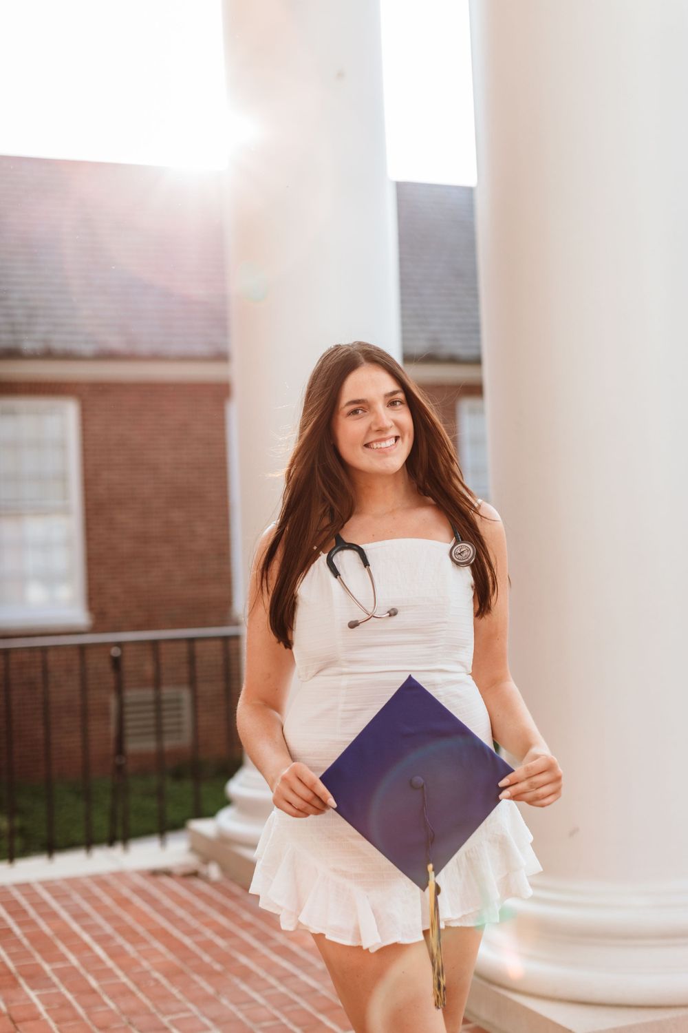 A student in a white dress holds a purple graduation cap while standing on a brick walkway near a brick building.