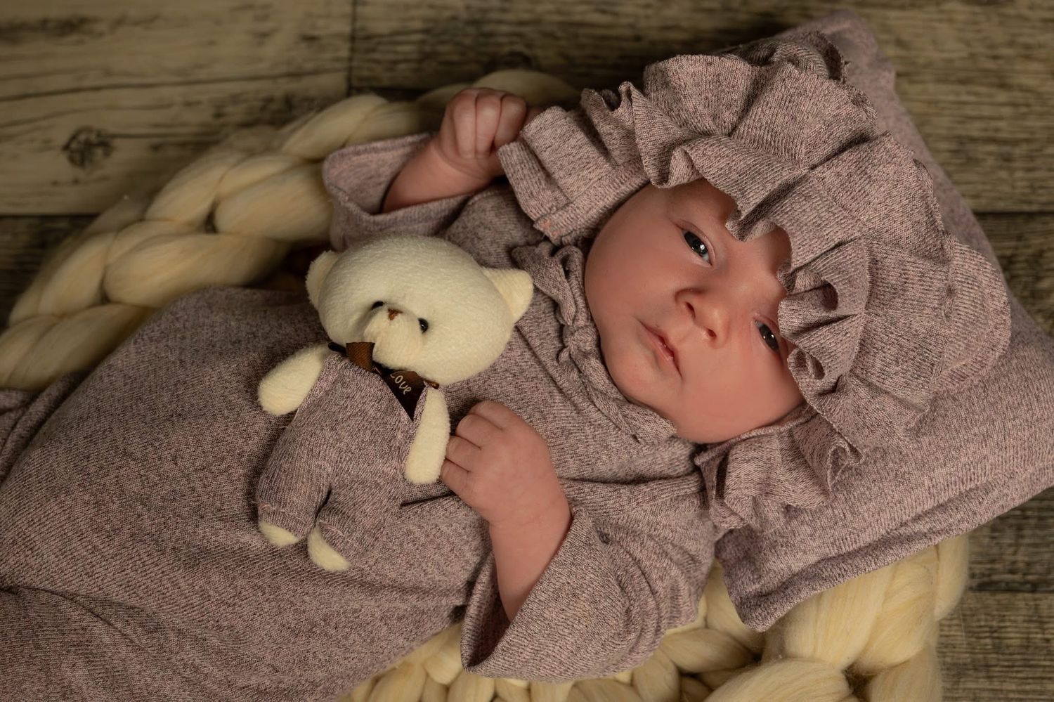 Adorable newborn baby in lavender outfit and bonnet holding a soft white teddy bear on cozy blanket.