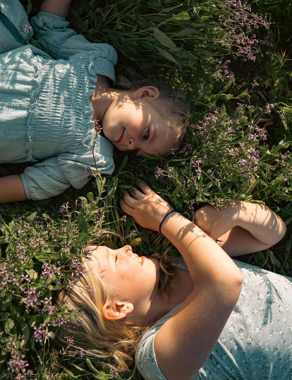 A mother and daughter lie together in a field of wildflowers, smiling at each other.