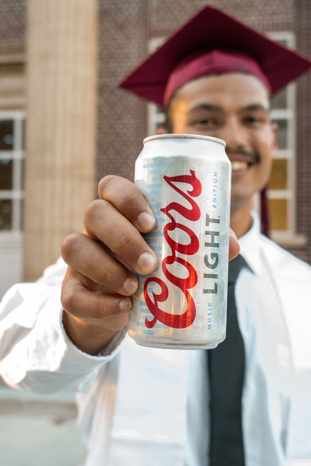 Smiling graduate in cap and gown holding a Coors Light Music Edition beer can toward the camera outdoors.
