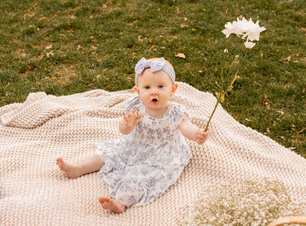 Baby girl in floral dress and bow headband sits on knit blanket outdoors, holding a white flower.