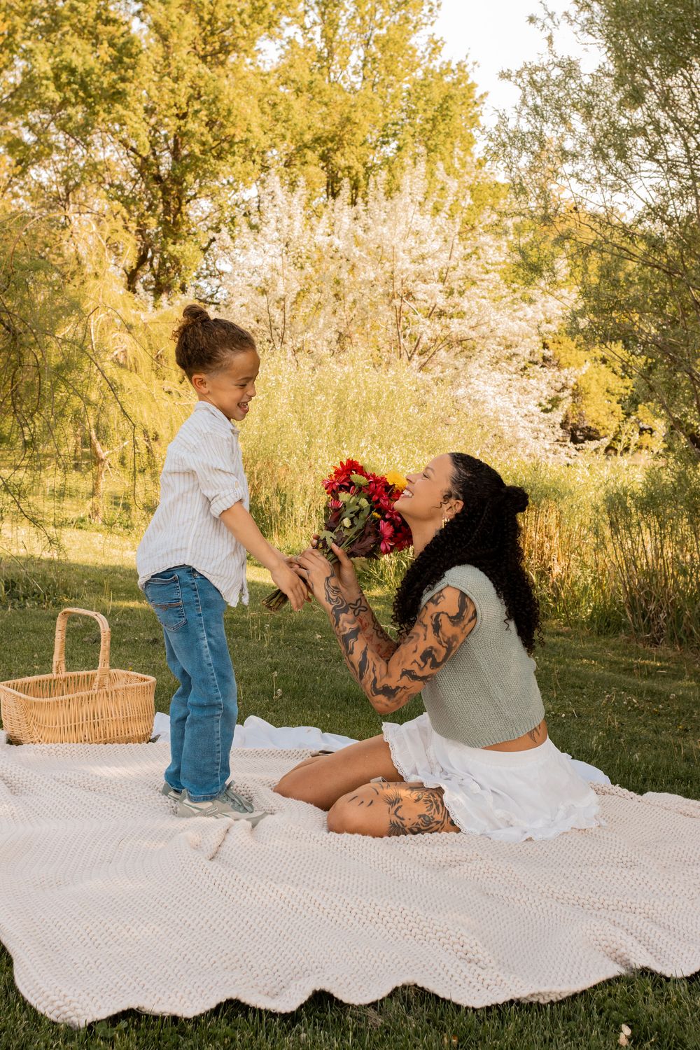 A tattooed mother receives a bouquet of flowers from her young child during a sunny outdoor picnic on a white blanket.