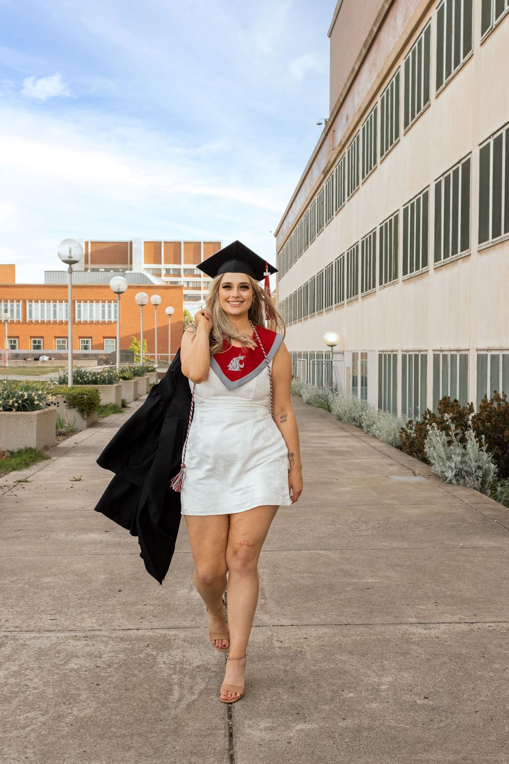 Young woman in white dress and graduation cap walks confidently outdoors, holding black gown, wearing WSU stole.