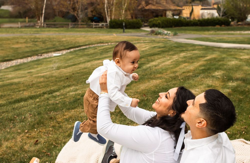 Happy family sitting on grass, parents lifting smiling baby outdoors in a sunny green park setting.
