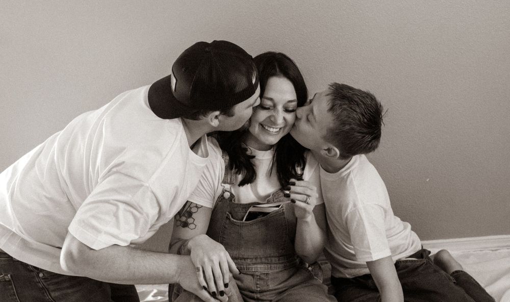 Two people kissing a smiling woman on the cheeks in a warm, joyful black and white portrait photo.