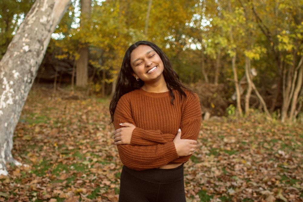 Young woman in a brown sweater smiling with arms crossed among autumn leaves in a park.