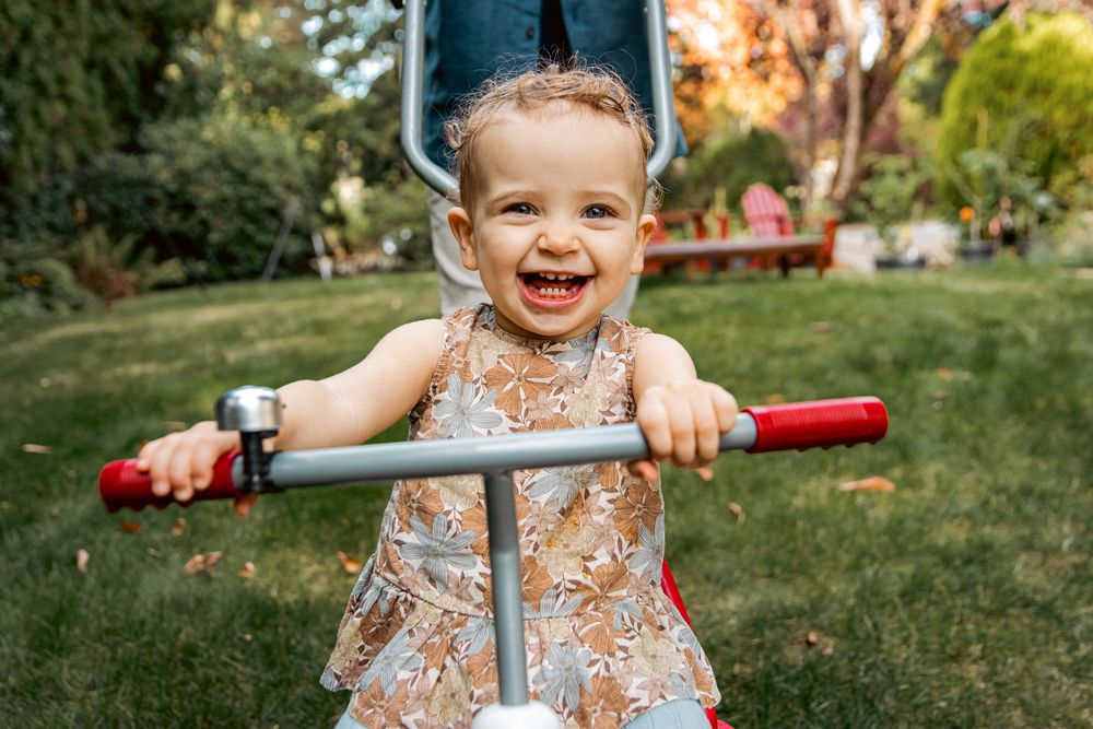Adorable laughing baby girl rides a small bike on green grass in a sunny park.