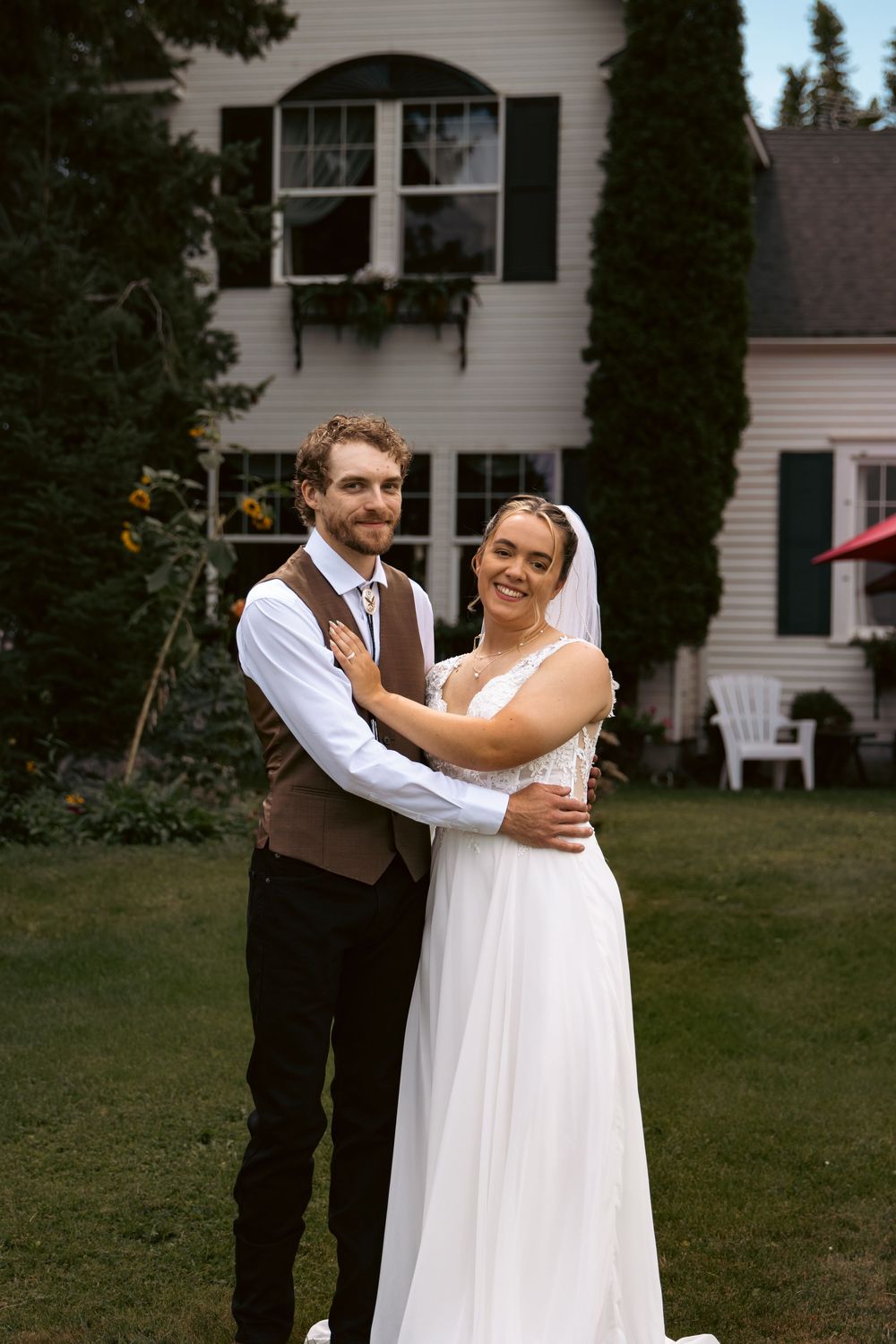 A smiling bride and groom embrace in front of a white farmhouse on their wedding day.