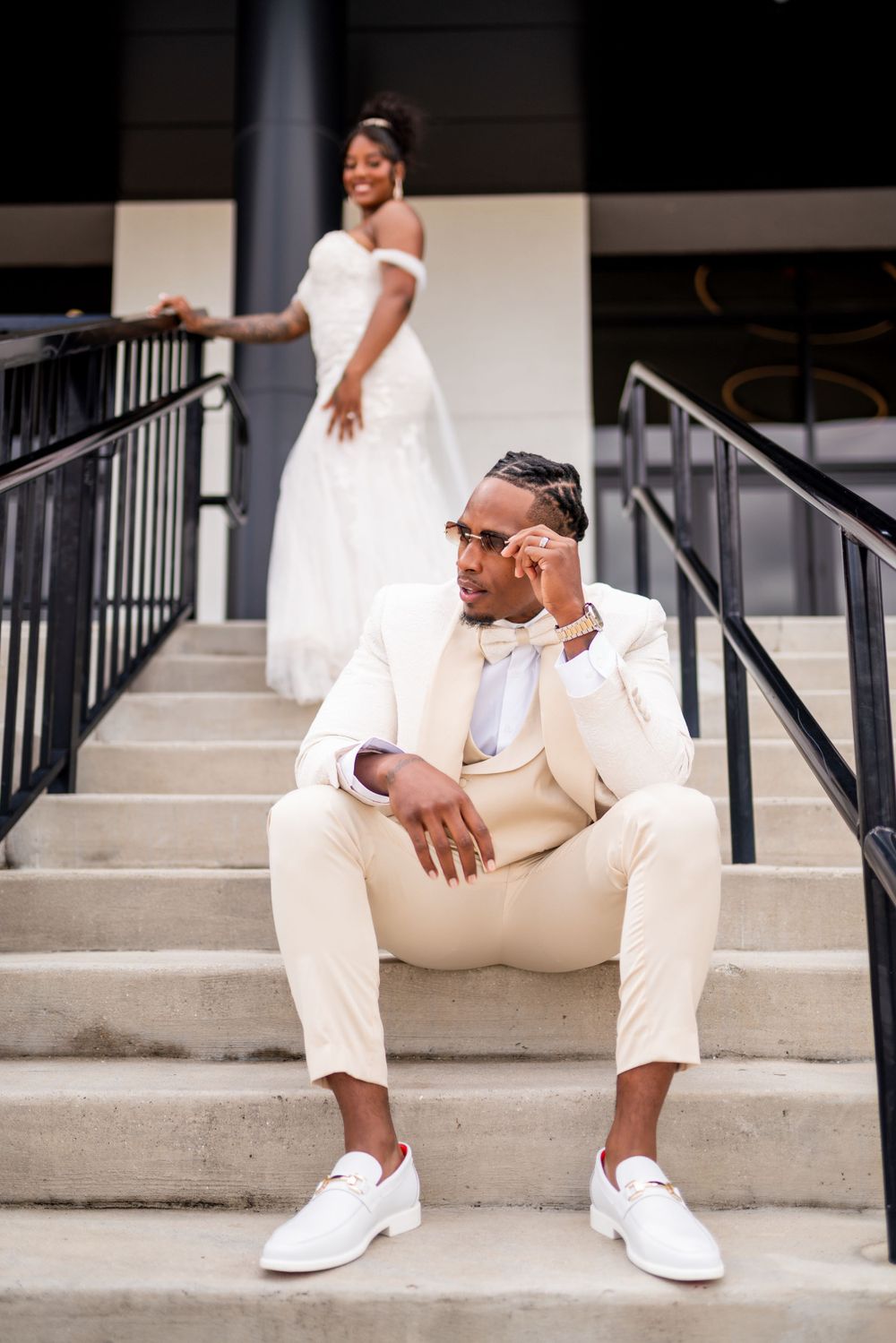 Bride and groom in stylish wedding attire pose on concrete steps, with groom in cream suit and white sneakers.