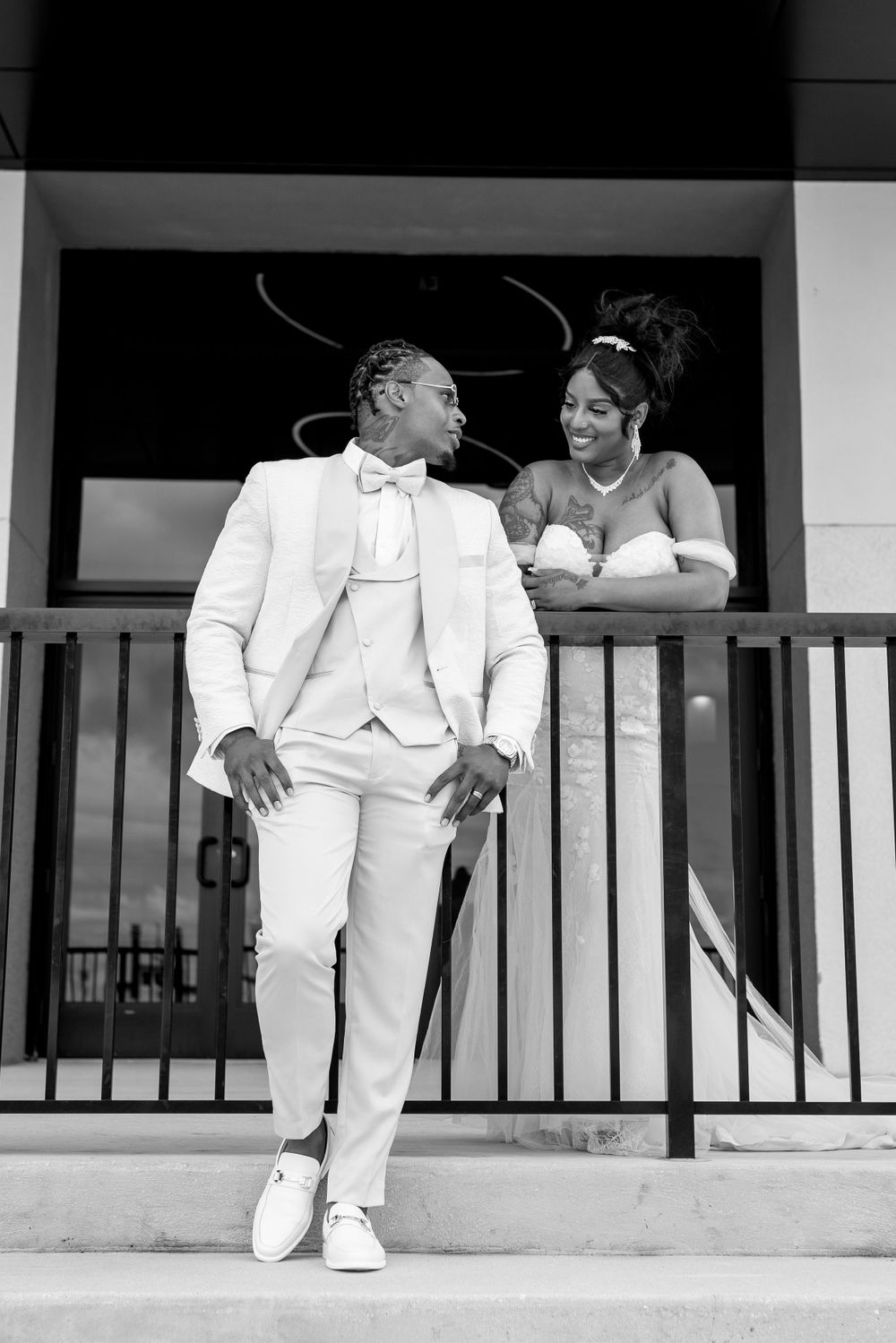A black and white photograph of someone in an all-white formal suit standing near a balcony railing while conversing.
