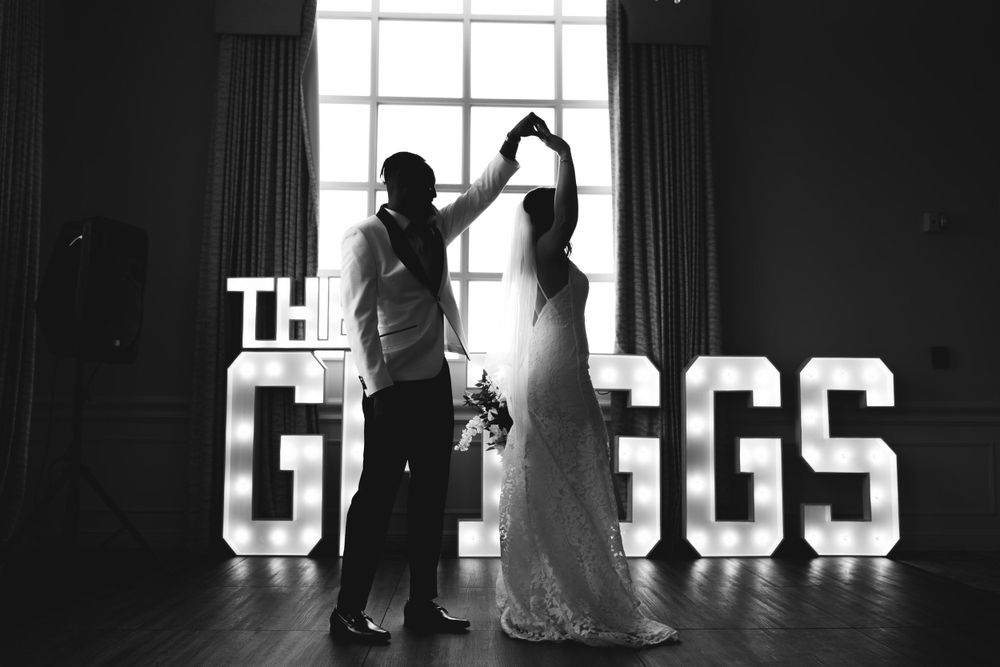 Black and white photo of a couple dancing in front of illuminated GREGGS marquee letters at their wedding reception.