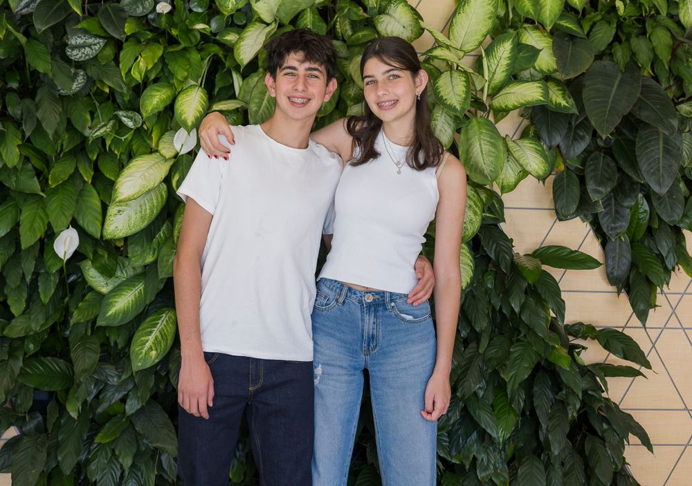 Two young people in casual white t-shirts pose against a leafy green wall background.