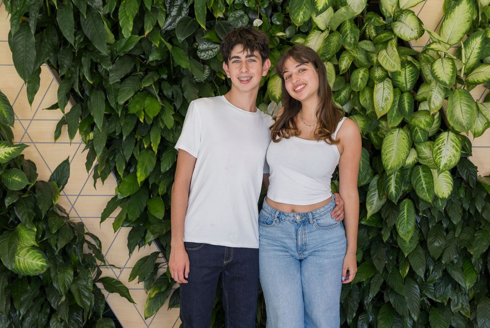 Two people in white tops and jeans pose casually against a wall covered in green foliage.