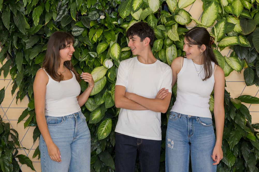 Three people in casual white tops and jeans share a laugh against a green leafy backdrop.