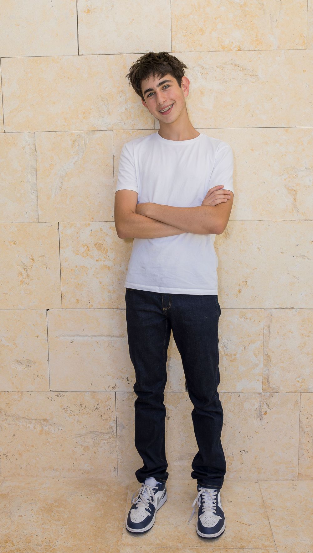 A person in a white t-shirt and dark jeans poses with arms crossed against a stone wall.