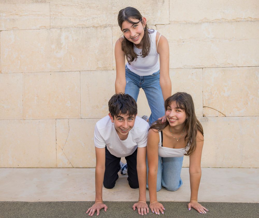 Three people in casual attire create a playful human pyramid pose against a stone wall.