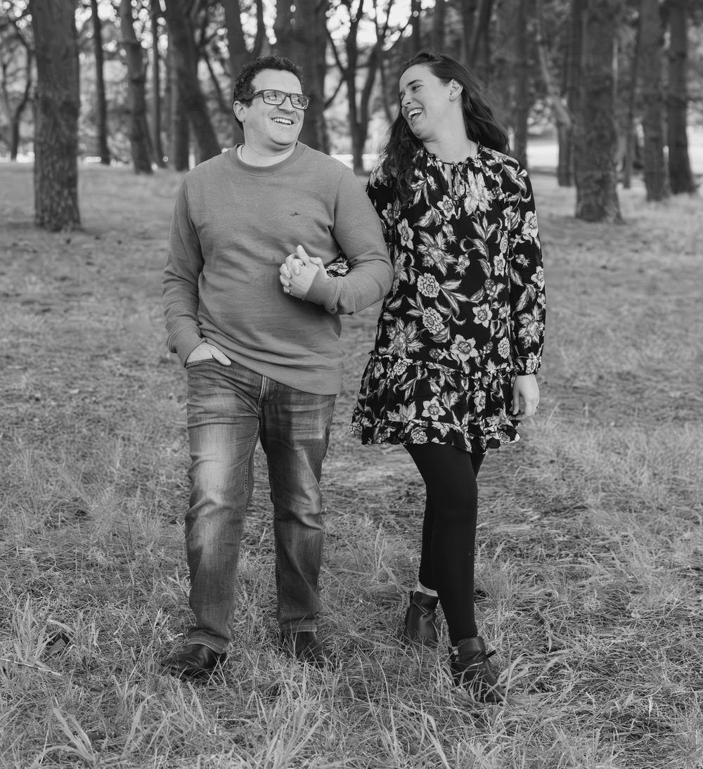 A couple poses together in black and white photography among pine trees in a forest setting.
