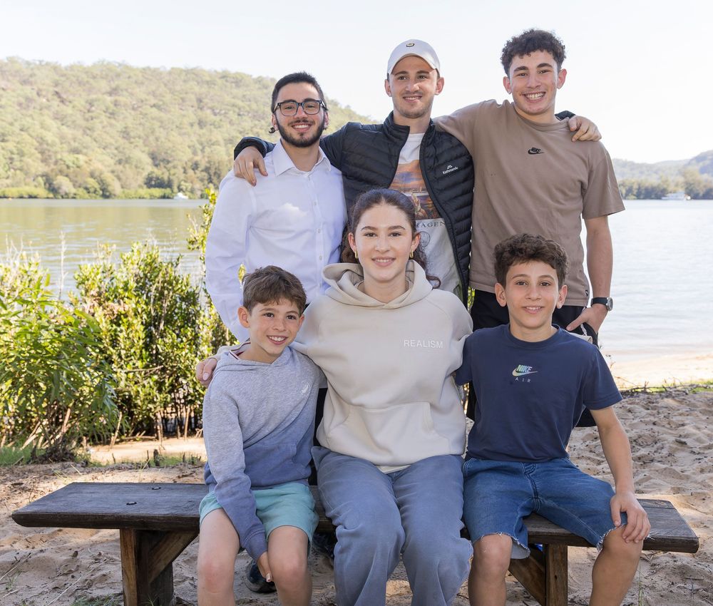 Group enjoying a sunny day together sitting on a bench by a lake with trees and mountains in the background.
