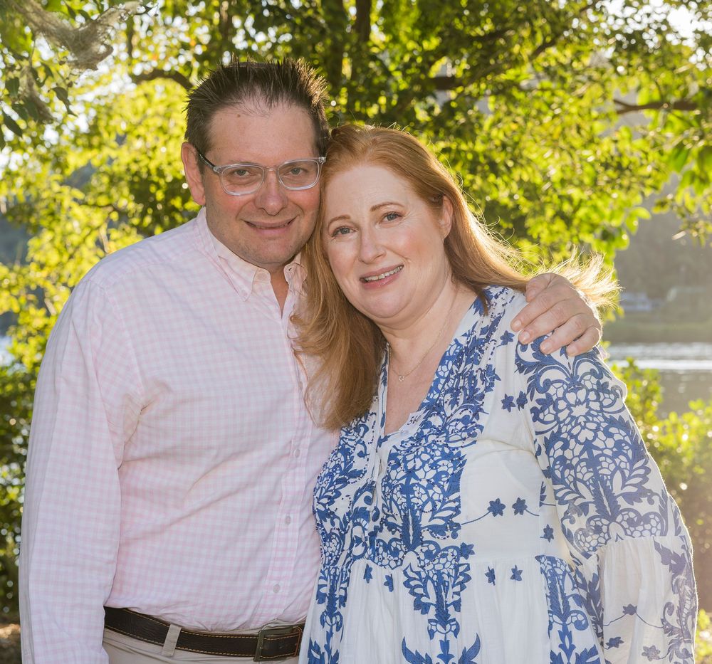 Couple smiling together outdoors on a sunny day with green foliage in the background.