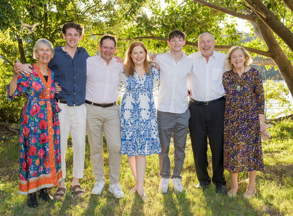 Group in casual attire posing outdoors together under a tree on a sunny day.