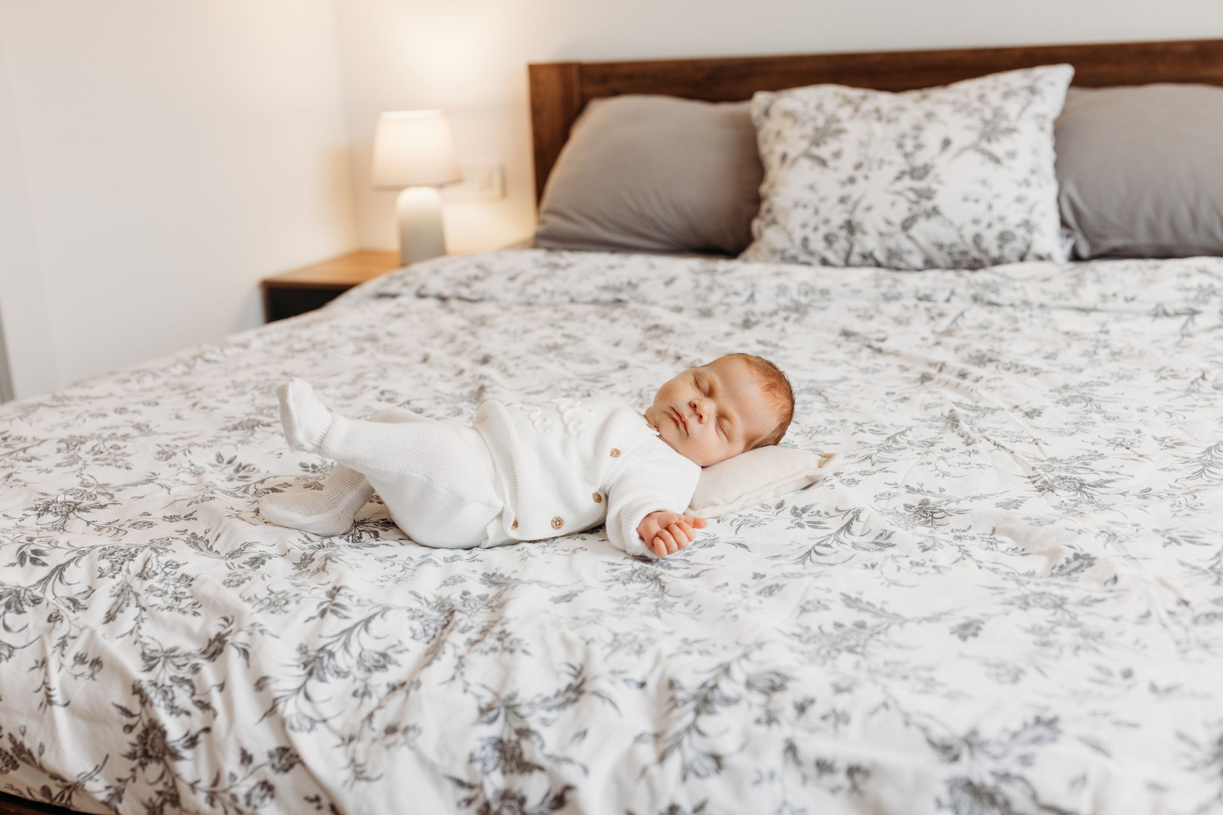 A baby in white sleepwear lies on a gray and white floral patterned bed.
