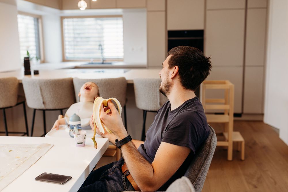 Person in a dark t-shirt sits at a wooden table in a bright minimalist kitchen, making hand gestures.