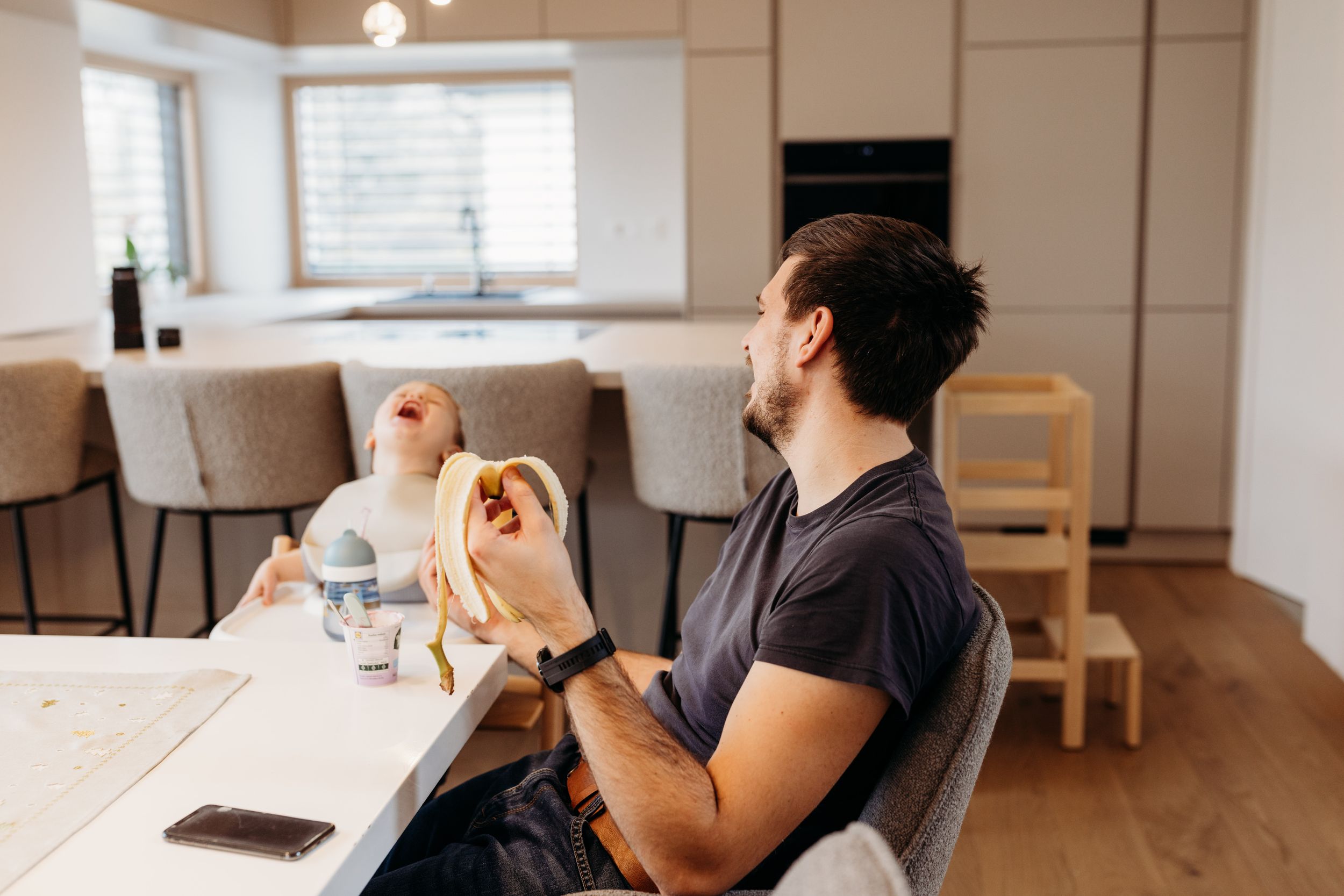 Person in a dark t-shirt sits at a wooden table in a bright minimalist kitchen, making hand gestures.