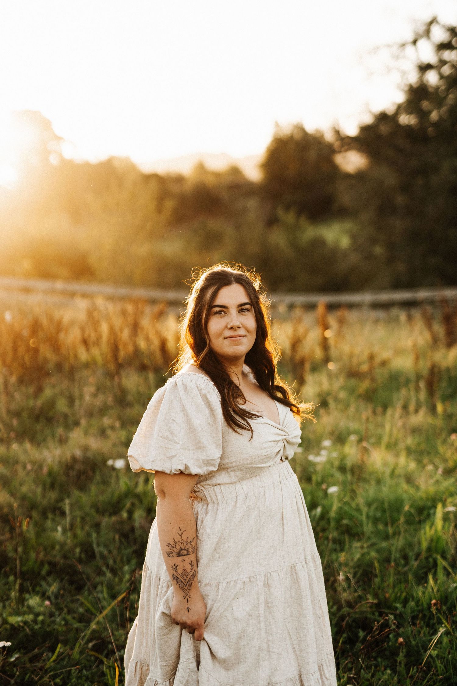 Glowing sunset maternity portrait of woman in white dress standing in golden field.