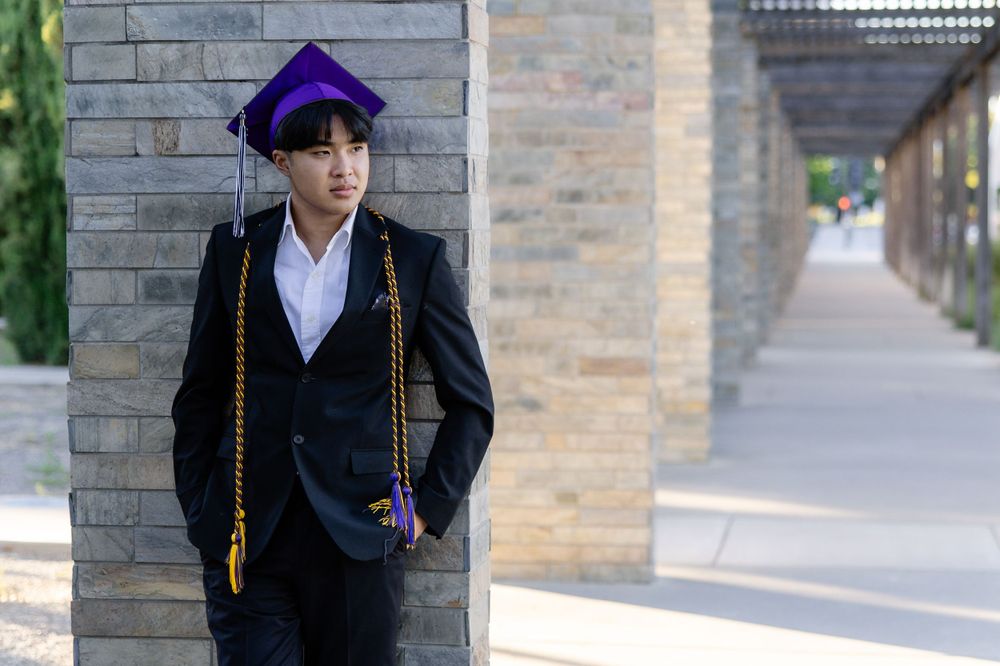 Graduate in purple cap and black gown poses against brick wall with graduation cords.