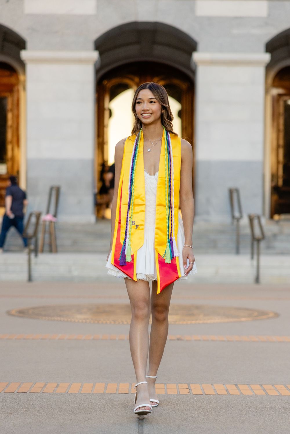 Graduate in white dress with colorful graduation stole standing in front of arched building.