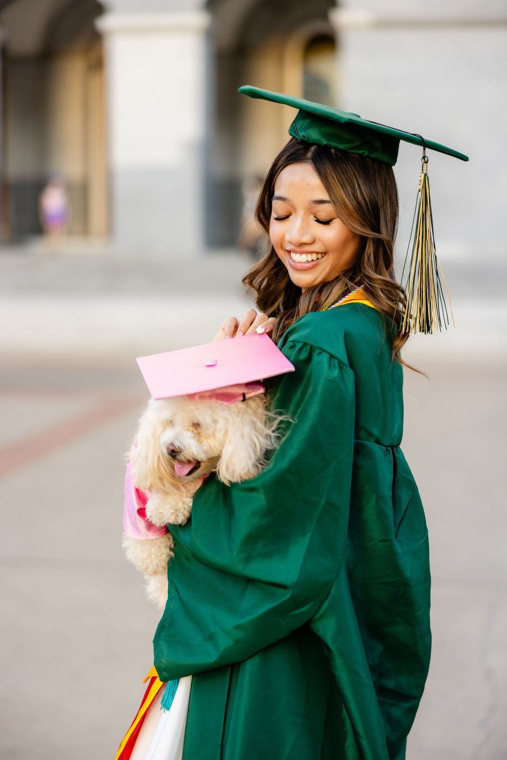Graduate in green cap and gown holds small white dog wearing pink graduation cap.