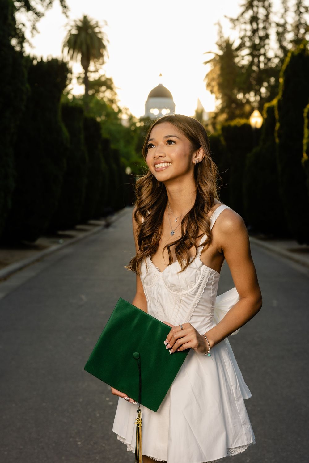 Graduate in white dress holding green folder on palm tree-lined path at sunset.