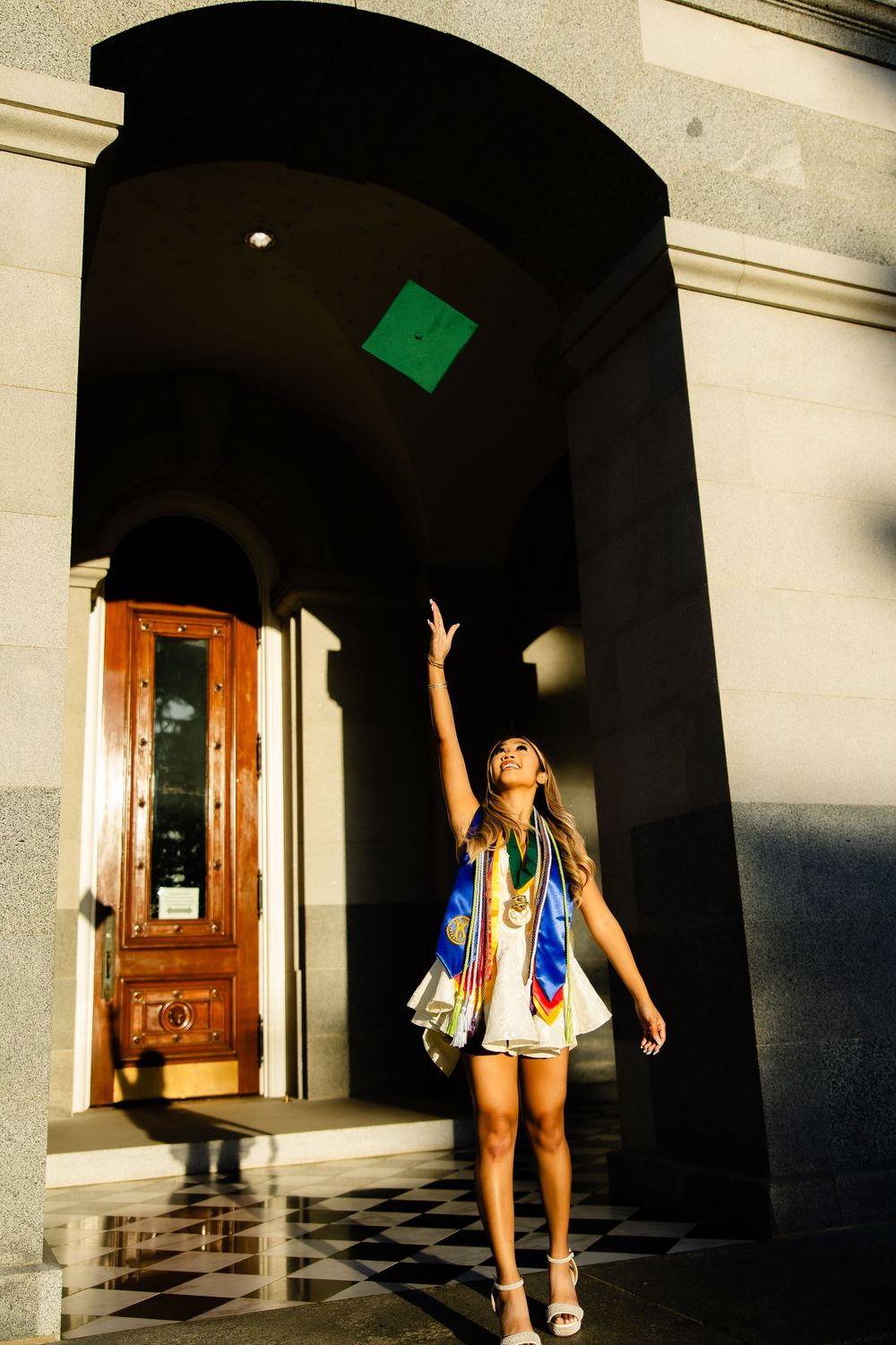 Graduate in academic dress reaching up toward green sign in arched doorway.