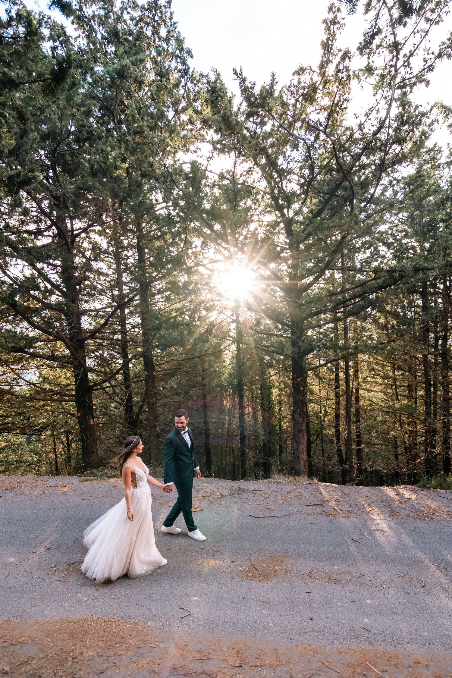 Wedding couple walking on forest path with sunlight streaming through evergreen trees.