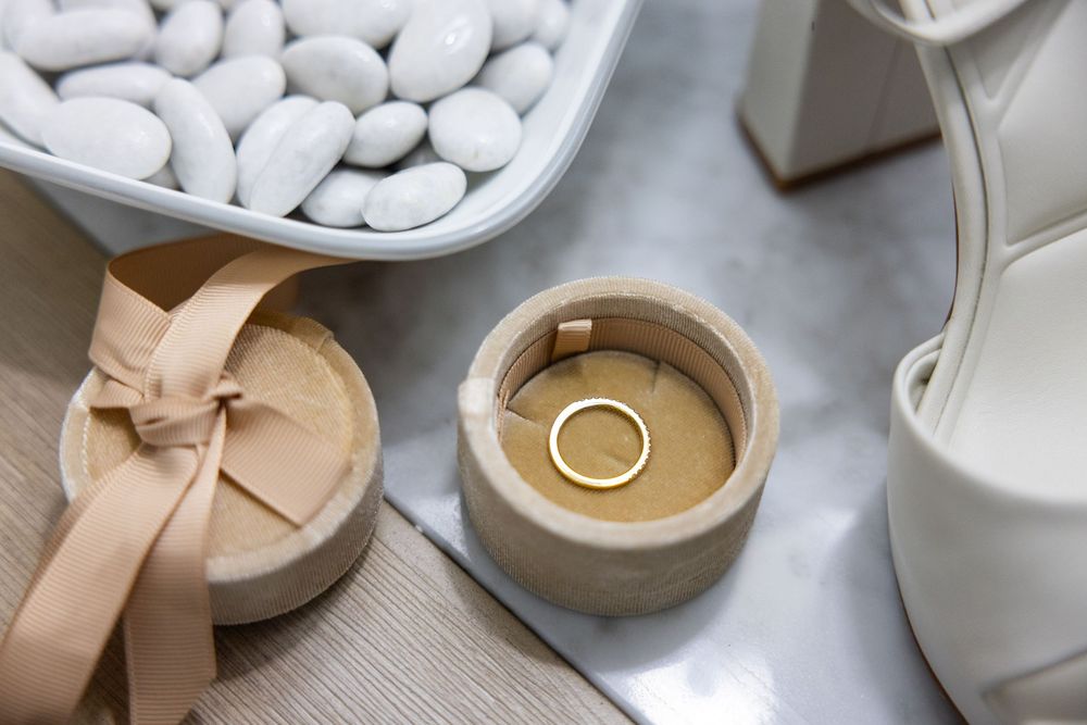 A gold wedding ring in a round wooden box surrounded by white stones and bridal accessories.