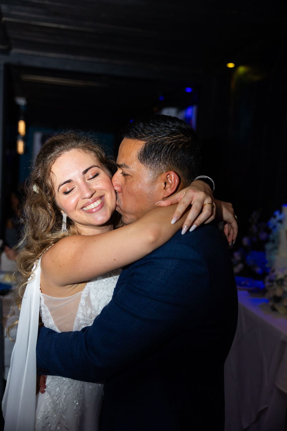 A smiling bride embraces her groom as he kisses her cheek during a romantic wedding reception.